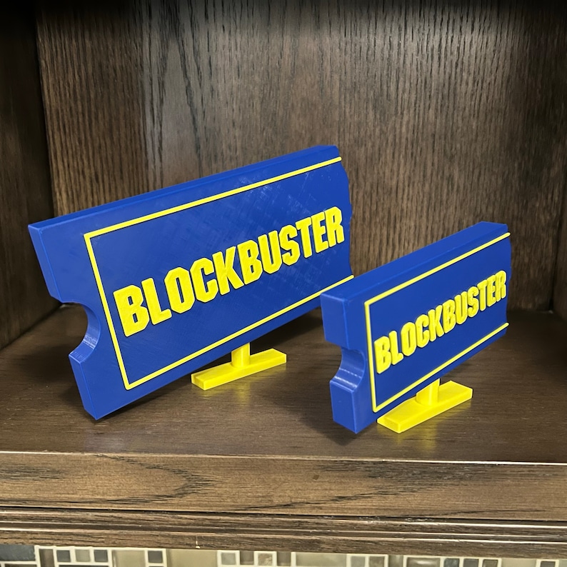 May include: Two blue and yellow signs with the word "Blockbuster" printed on them. The signs are made of plastic and are propped up on small stands.