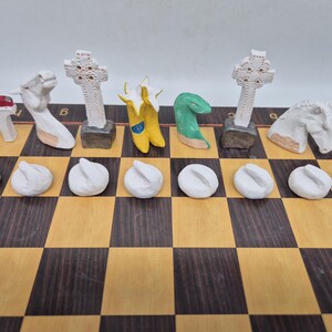 May include: A set of custom chess pieces on a wooden board. The back row includes white and colored figures shaped like a red-topped arch, a horse head, a Celtic cross, yellow boots, a green snake, another Celtic cross, and a horse head. Eight white pawns.