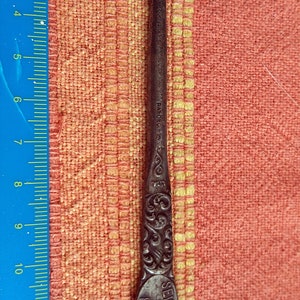 Könnte beinhalten: Ein dunkler Metall-Knopfhaken mit einer Länge von etwa 14,5 Zentimetern. Der Griff ist mit einem verzierten Blumenmuster und einer ovalen Plakette mit der Aufschrift SEVEN SISTERS 53 & 55 ROAD versehen. Ein blaues Lineal liegt daneben.
