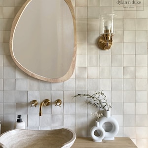 May include: A bathroom vanity with a white ceramic sink, gold faucets, and a wooden, oval-shaped mirror. The wall is tiled with white square tiles. There are two white ceramic vases with a ring design on the counter, one with a sprig of greenery.