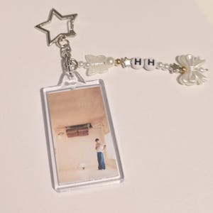 May include: A clear acrylic keychain with a rectangular photo of a person in a room. The keychain has a silver star-shaped charm and a beaded strand with the letters "HH". Decorative white butterfly and bow charms complete the design.