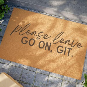 May include: A rectangular doormat with a tan surface and dark gray text. The text reads "please leave GO ON, GIT." The mat is placed on a gray stone surface, with greenery visible in the corners.