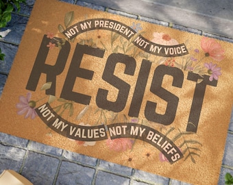 May include: A rectangular doormat with the word "RESIST" in large, dark brown letters. The mat is tan with floral accents and the phrases "NOT MY PRESIDENT," "NOT MY VOICE," "NOT MY VALUES," and "NOT MY BELIEFS."