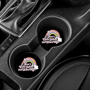 May include: Two black car coasters with a rainbow and cloud design. The text on the coasters reads "I Don't Care What the Bible Says".