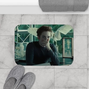 Puede incluir: Una alfombra de baño rectangular con una escena de película. La alfombra muestra a una persona con una parte superior oscura, con una ventana y una imagen enmarcada en el fondo. Dos zapatillas de punto gris están en primer plano.