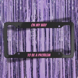 Op de afbeelding: Zwart kentekenplaatraam met de tekst "ON MY WAY TO BE A PROBLEM" in roze. Het frame is rechthoekig met afgeronde hoeken en staat tegen een achtergrond van verticale paarse slingers.