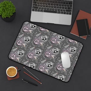 May include: Gray mousepad with a repeating pattern of skulls, axes, and knives. The skulls have the text "THE HUSBAND DID IT" and "STAY FOREST". A computer mouse sits on the pad, with a laptop and other office supplies in the background.
