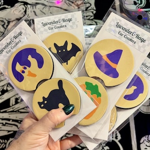 May include: A collection of car coasters with Halloween-themed designs. Each coaster features a beige background with a black border and a different graphic: a ghost, a bat, a witch's hat, a black cat, and a taco. The text "Lavender & Rage Car Coasters" is printed on the packaging.