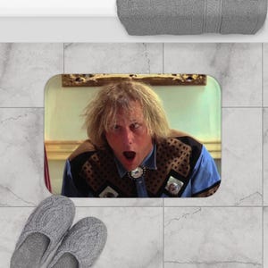 Puede incluir: Alfombra de baño rectangular con un hombre sorprendido con cabello rubio y una camisa azul. La alfombra tiene un borde verde claro y una foto del hombre. Zapatillas grises en primer plano.