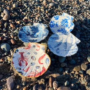 Può includere: Quattro conchiglie di capasanta decorative con disegni dipinti. Una conchiglia presenta un motivo a onde blu, un'altra ha un design floreale blu e bianco, una terza mostra un motivo di aragosta e conchiglie, e l'ultima ha un motivo di pesce blu. Le conchiglie sono disposte su una superficie di ciottoli.