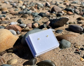 Puede incluir: Un par de pendientes de botón pequeños y redondos de piedras preciosas amarillas presentados en una caja de joyería blanca abierta. La caja está sobre una playa arenosa con varias rocas y guijarros. El fondo muestra el mar y un día soleado.