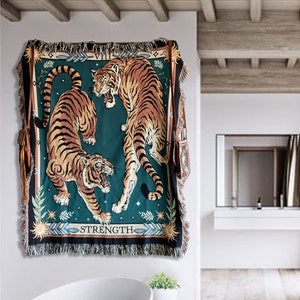 May include: A tapestry wall hanging with a green background featuring two tigers. The tigers are facing each other, one is standing on its hind legs and the other is lying down. The tapestry has a fringe border and the text "STRENGTH" is printed at the bottom.