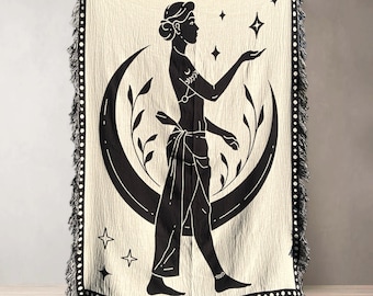 May include: A woven throw blanket with a cream background and a black silhouette of a woman standing in front of a crescent moon. The woman is reaching out to touch a star. The design is surrounded by black leaves and stars.