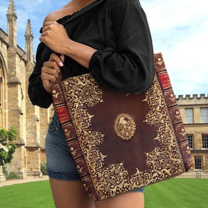 Op de afbeelding: Een bruine tote bag met een gouden, sierlijk ontwerp. De tas is gemaakt om eruit te zien als een oud boek met een gouden rug en gouden letters. De tas wordt gedragen door iemand die een zwart shirt en een blauwe spijkerbroek draagt.
