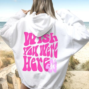 Op de afbeelding: Witte hoodie met een roze "Wish You Were Here" afbeelding en een roze bloem op de rug.