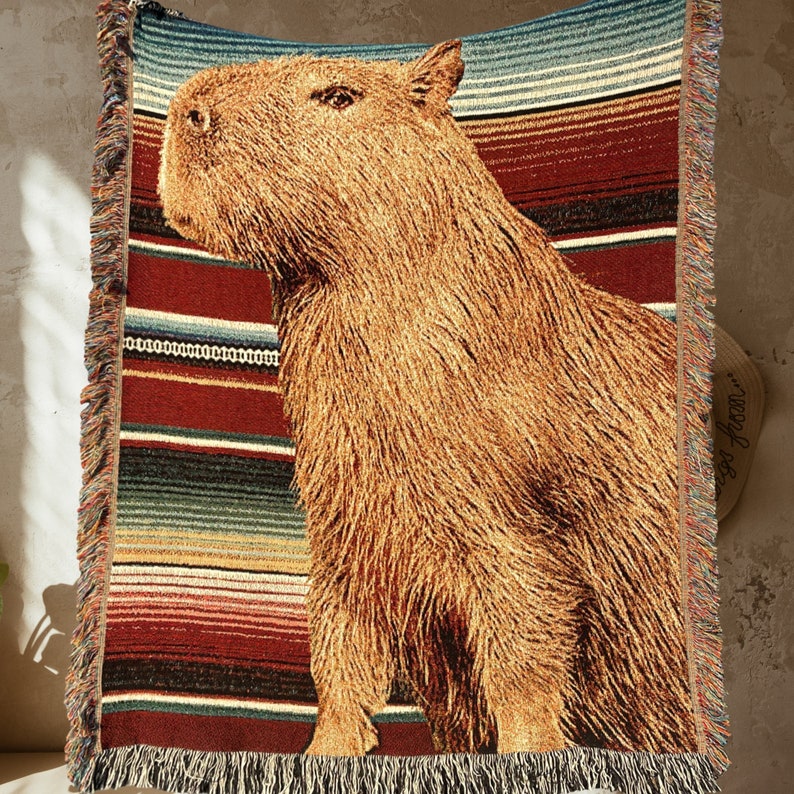 The ideal capybara gift, a capybara blanket! This capybara woven blanket with fringe is all cotton. The background has a vintage Mexican blanket aesthetic with a large, central capybara front and center. All cotton original design. Made in the US.