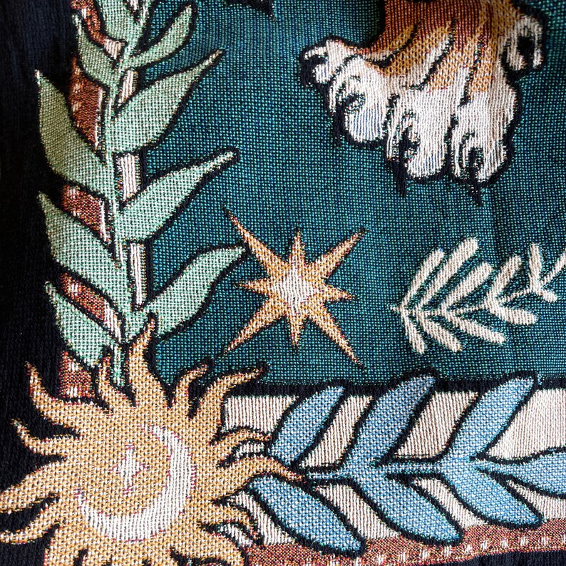 May include: A tapestry with a blue background featuring a stylized sun, a star, and a leafy branch. The sun is gold and has a crescent moon inside. The star is gold with a white center. The branch is green and white.