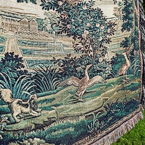 May include: A woven tapestry throw blanket featuring a detailed landscape scene with a large tree, a fountain, a stone building, a dog, and a goose with wings spread. The textile displays a palette of deep greens, browns, and cream tones with fringe.