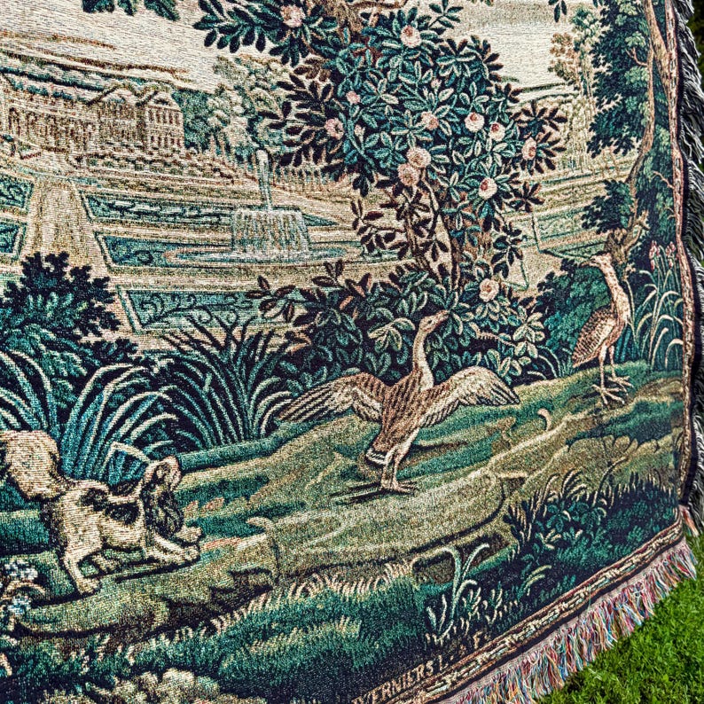 May include: A woven tapestry throw blanket featuring a detailed landscape scene with a large tree, a fountain, a stone building, a dog, and a goose with wings spread. The textile displays a palette of deep greens, browns, and cream tones with fringe.