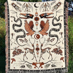 May include: A woven tapestry throw blanket with a cream background and a symmetrical design featuring a variety of creatures, including snakes, owls, deer, and insects. The design is in shades of brown, black, and red.