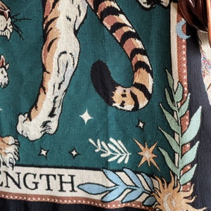 May include: A tapestry featuring a tiger with a green background and gold stars. The tiger's tail is curled around its body. The tapestry has the word "STRENGTH" at the bottom.