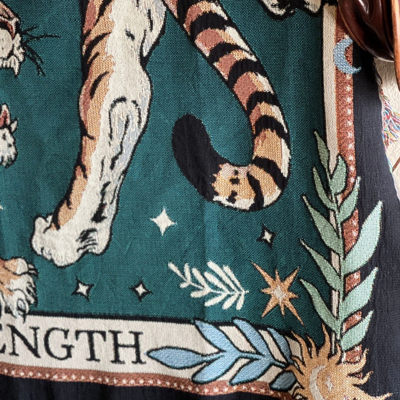 May include: A tapestry featuring a tiger with a green background and gold stars. The tiger's tail is curled around its body. The tapestry has the word "STRENGTH" at the bottom.
