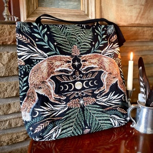 A black tote bag with a tapestry design featuring two foxes facing each other in a forest scene. The foxes are orange and brown with white accents. The forest scene is filled with green leaves, ferns, and a crescent moon with stars. The bag has a black strap and is made of a woven fabric.
