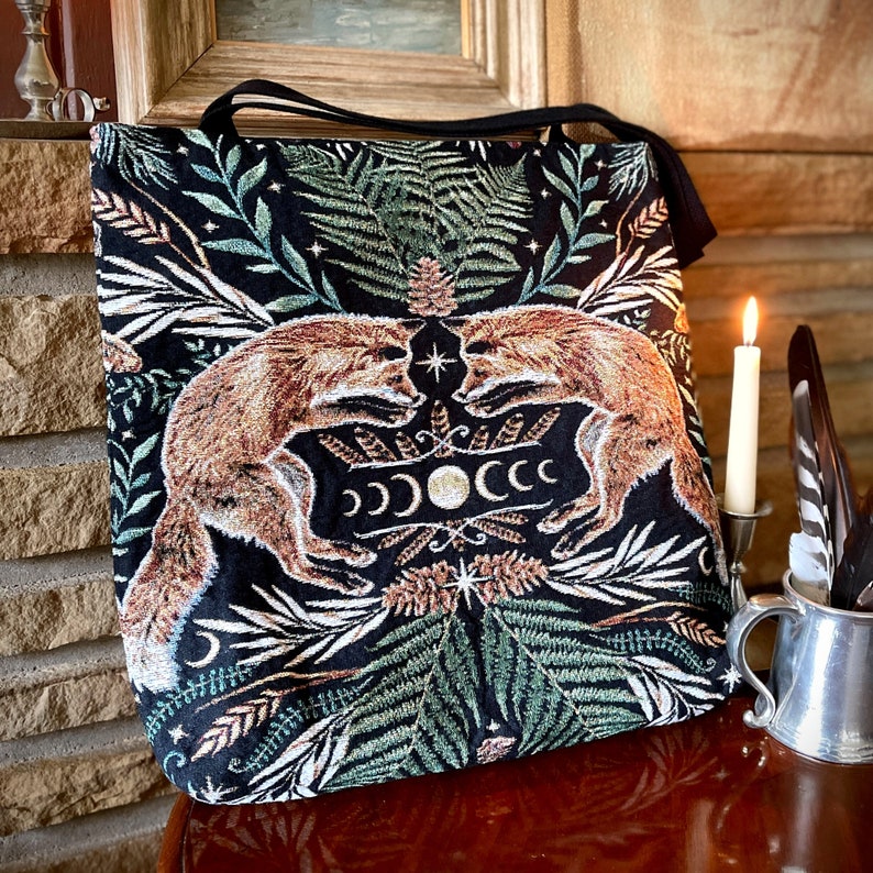 A black tote bag with a tapestry design featuring two foxes facing each other in a forest scene. The foxes are orange and brown with white accents. The forest scene is filled with green leaves, ferns, and a crescent moon with stars. The bag has a black strap and is made of a woven fabric.