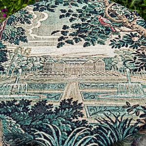 May include: A round woven tapestry featuring a detailed landscape scene with a large manor house, formal gardens, fountains, and trees in shades of deep green, teal, beige, and brown. A small bird is perched on a branch in the upper right corner.