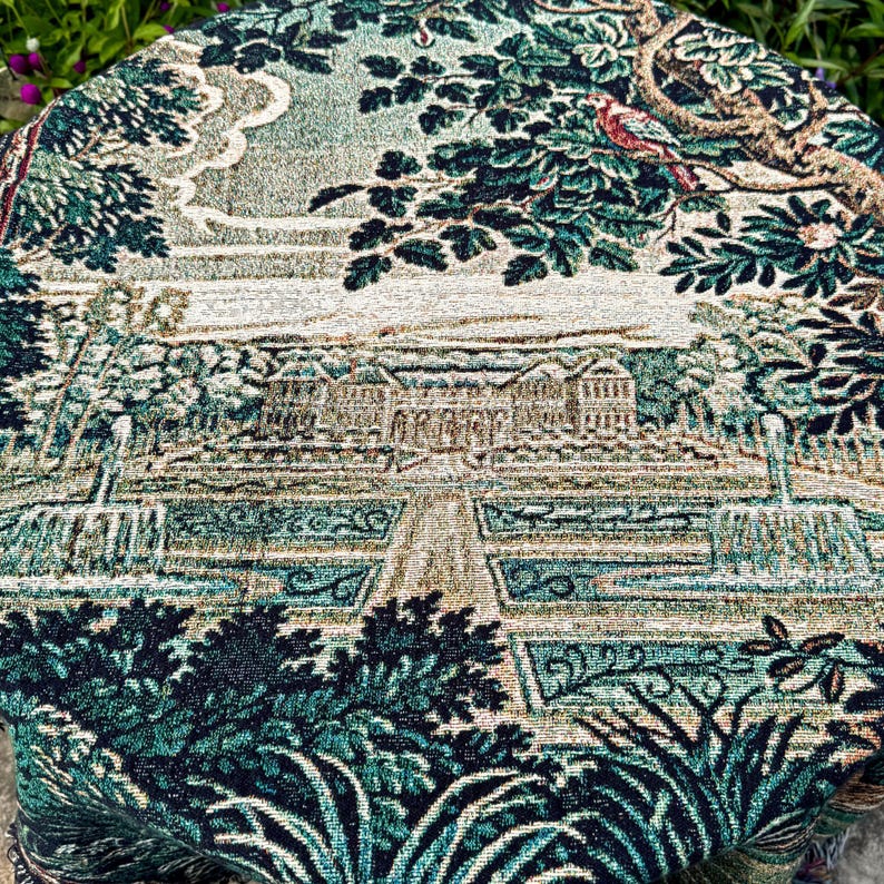 May include: A round woven tapestry featuring a detailed landscape scene with a large manor house, formal gardens, fountains, and trees in shades of deep green, teal, beige, and brown. A small bird is perched on a branch in the upper right corner.