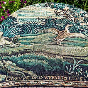 May include: A round woven tapestry featuring a detailed scene of a bird with outstretched wings and a dog in a lush landscape. The design includes deep shades of green, blue, and tan, with the text LAVEVVE DEG WERNIERS L. F. printed at the bottom.