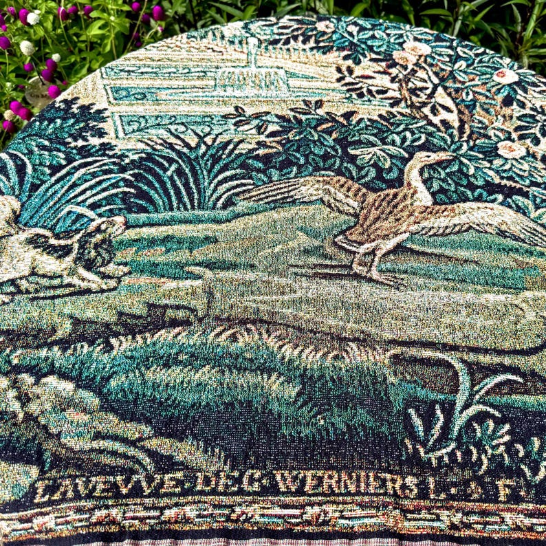 May include: A round woven tapestry featuring a detailed scene of a bird with outstretched wings and a dog in a lush landscape. The design includes deep shades of green, blue, and tan, with the text LAVEVVE DEG WERNIERS L. F. printed at the bottom.