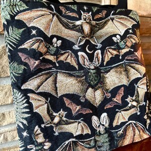 May include: Black tote bag with a tapestry design featuring bats and a crescent moon. The bats are in various poses and sizes, with some having a more detailed design than others.