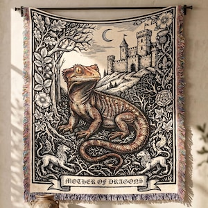 May include: A tapestry featuring a detailed illustration of a lizard with a castle in the background. The artwork is in black and white with a brown lizard, and the words "MOTHER OF DRAGONS" are displayed on a banner.