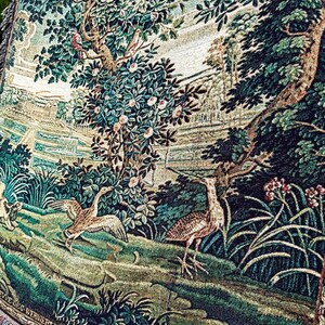 May include: A woven tapestry featuring a landscape scene with two birds in the foreground, one with wings spread and one standing. The background displays lush green trees, flowering bushes, and a distant architectural structure with a fountain.