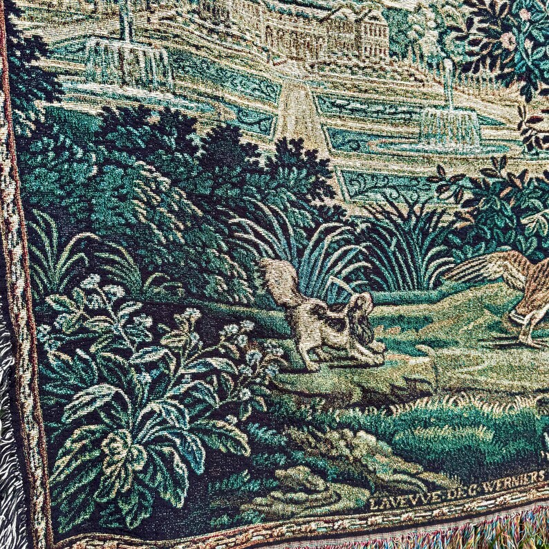 May include: A woven tapestry featuring a garden scene with fountains, lush green foliage, a small dog, and a bird. The lower right corner displays the text LAVEVVE DE G WERNIERS. The fabric has a textured, multi-colored thread construction.