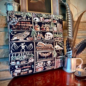 Puede incluir: Un bolso tote azul oscuro con un dise&ntilde;o de tapiz que presenta un b&uacute;ho, una calavera, libros y las palabras "Herbal Apothecary". La bolsa tiene un patr&oacute;n de cuadr&iacute;cula con varias ilustraciones de tem&aacute;tica oculta. Una taza de metal con plumas est&aacute; a la derecha.