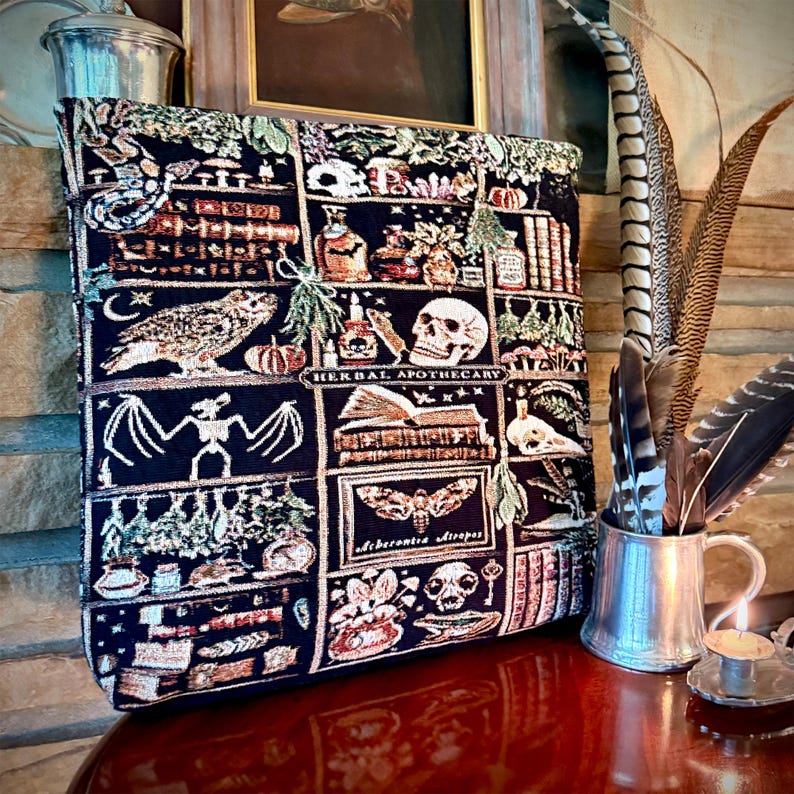 Puede incluir: Un bolso tote azul oscuro con un dise&ntilde;o de tapiz que presenta un b&uacute;ho, una calavera, libros y las palabras "Herbal Apothecary". La bolsa tiene un patr&oacute;n de cuadr&iacute;cula con varias ilustraciones de tem&aacute;tica oculta. Una taza de metal con plumas est&aacute; a la derecha.