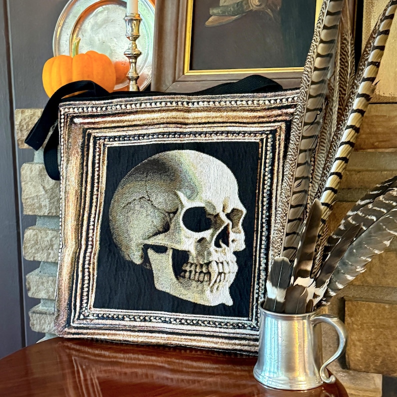 K&ouml;nnte beinhalten: Eine schwarze Stofftasche mit einem goldenen Rahmen, der einen detailliert gestickten Totenkopf zeigt. Die Tasche hat einen langen Riemen und steht auf einem Holztisch.