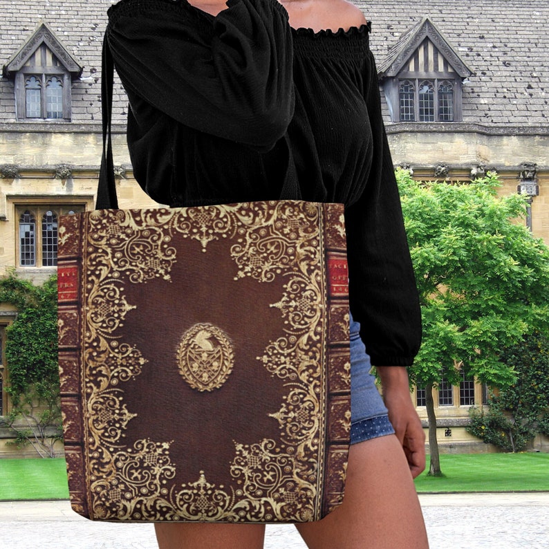 Op de afbeelding: Een bruine tote bag met een vintage boekomslagontwerp. De omslag heeft ingewikkelde gouden filigraan en een wapenschild met een vogel erin. De tas heeft een lange band en wordt door iemand gedragen.