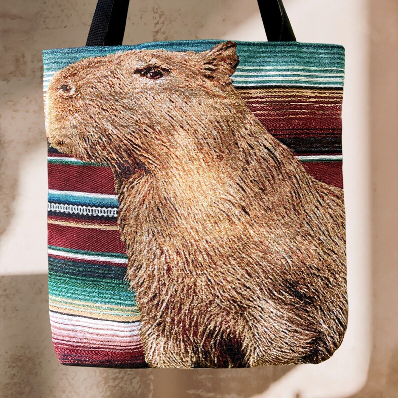 May include: A brown and white capybara tote bag with a colourful striped pattern. The bag has a brown leather handle and is made of a woven fabric.