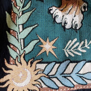 Pode incluir: Uma tape&ccedil;aria com fundo azul que apresenta um sol estilizado, uma estrela e um ramo frondoso. O sol &eacute; dourado e tem uma lua crescente dentro. A estrela &eacute; dourada com um centro branco. O ramo &eacute; verde e branco.