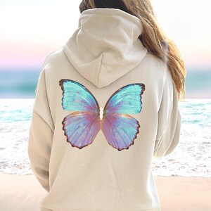 May include: A light beige hoodie with a large iridescent blue butterfly printed on the back. The butterfly has purple and blue wings with a brown outline.