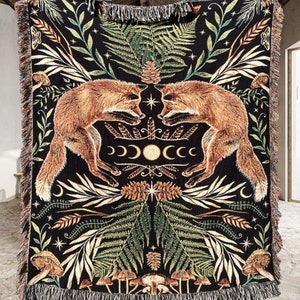 Fox woven blanket with crescent moons and botanicals. A black woven throw blanket with a tapestry design featuring two foxes facing each other in a forest scene. The foxes are orange and white, and the forest is filled with green ferns, leaves, and flowers. The blanket has a fringe border. 100% cotton & machine washable.