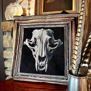 May include: A black and white tapestry depicting a skull illustration. The skull is the central focus, framed by a decorative border with gold and brown detailing. The tapestry is displayed on a wooden surface, with a metallic jug of feathers.