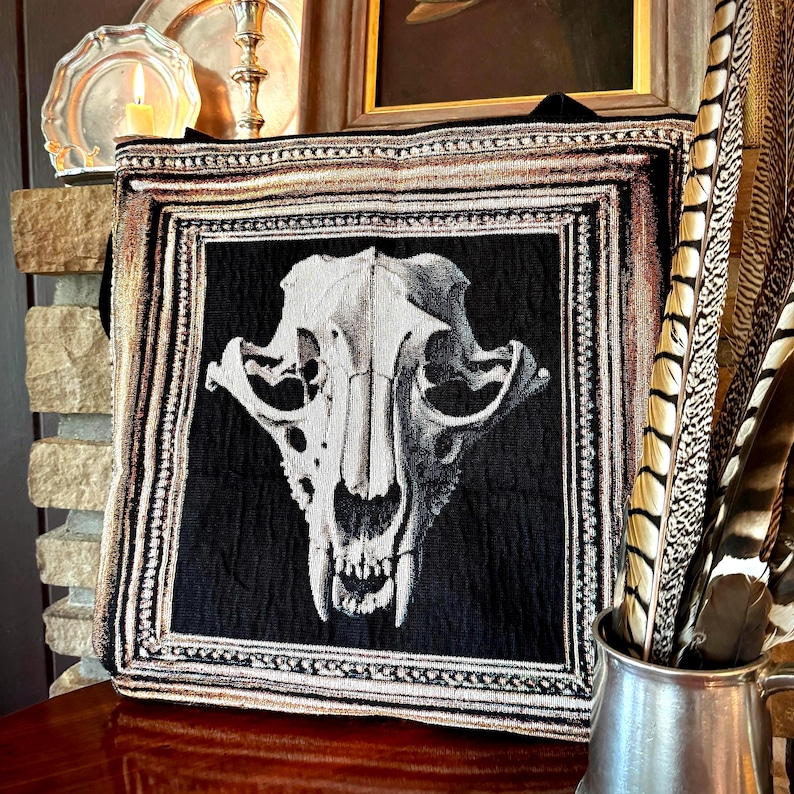May include: A black and white tapestry depicting a skull illustration. The skull is the central focus, framed by a decorative border with gold and brown detailing. The tapestry is displayed on a wooden surface, with a metallic jug of feathers.