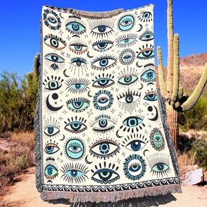 May include: A white woven throw blanket with a repeating pattern of blue and green eyes. The eyes are surrounded by black and white geometric designs.