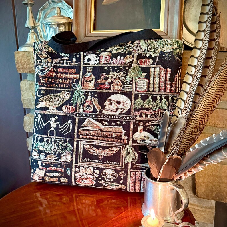 With a vintage Halloween aesthetic, this HERBAL APOTHECARY WOVEN TAPESTRY TOTE BAG has a witchy book nook aesthetic that will delight dark academia, vulture culture, oddities & whimsigoth lovers alike. Highly detailed apothecary bottles, mushrooms, stars, owls, skulls, hanging herbs, feather quills, snakes, crystals, a drunken mandrake with his bubbling tankard & more are expertly woven into every inch of this special woven tapestry tote bag.