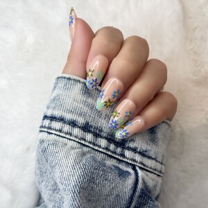 Peut inclure: Une main de mannequin portant de longs ongles en gel de forme amande, de couleur rose clair, avec un motif floral. Les fleurs sont bleues, vertes et jaunes.