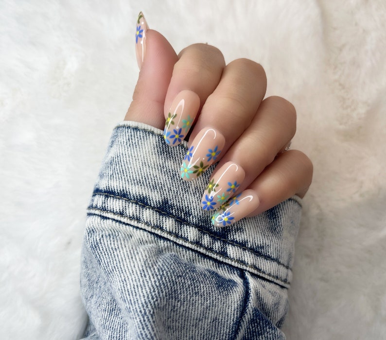 Peut inclure: Une main de mannequin portant de longs ongles en gel de forme amande, de couleur rose clair, avec un motif floral. Les fleurs sont bleues, vertes et jaunes.